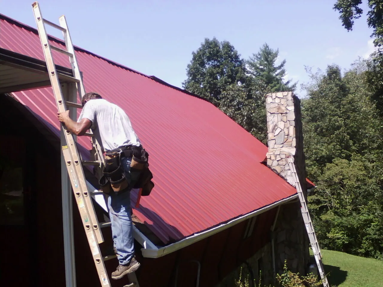 Roofer on a ladder beside a red metal roof and stone chimney — Soffit & Fascia Repair by Roofing Brothers in Boyden